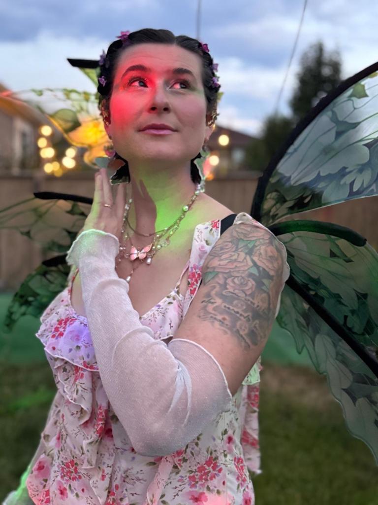 lauren, a white person, is wearing fairy wings, a pink and white dress, and white gloves. their brown hair is slicked down and has butterfly clips in it. they are looking up, their hand is near their face, and they are illuminated by green and red light.